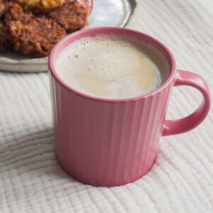 Chai Masala Latte in a pink cup with a plate of Pakoras with sauce on the side on a beige grey plate in the background