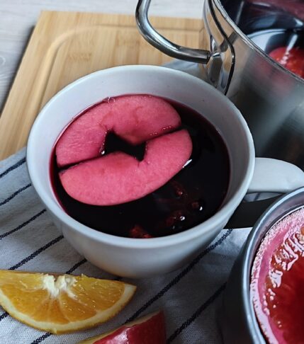 A full cup of Glühwein, with apple pieces floating in it and a pot of Glühwein can be seen in the background. Orange slices, and apple slices are scattered around on a striped cloth atop a working surface, serving as decoration.
