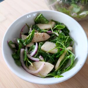 Apple Arugula Sumac Salad in a white salad bowl on a working surface with a large clear bowl in the background with the remaining portion of salad