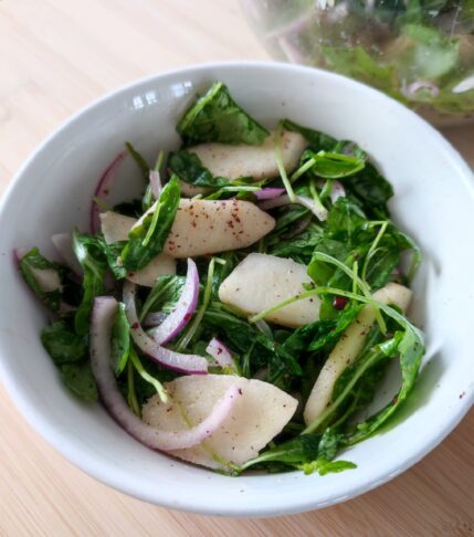 Apple Arugula Sumac Salad in a white salad bowl on a working surface with a large clear bowl in the background with the remaining portion of salad