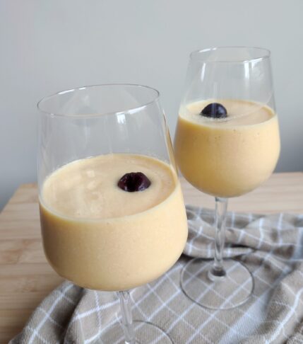 Rosé Mango Piña Colada in two wine glasses topped with one whole cherry on a warm taupe checkered fabric on a working surface