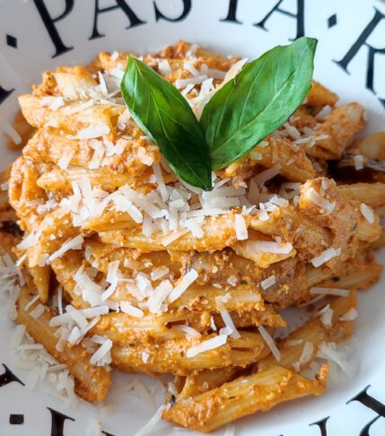 Pasta Pesto Rosso with Shiitake Mushrooms garnished with shredded parmigiano reggiano and basil leaves on a white plate with black lettering