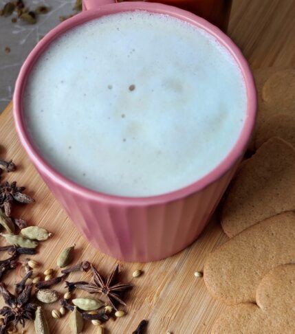 Masala Chai Latte in deep pink cup surounded with few hearts cookies on the right nd blend of whole spices on left and candle in a glass jar at the back