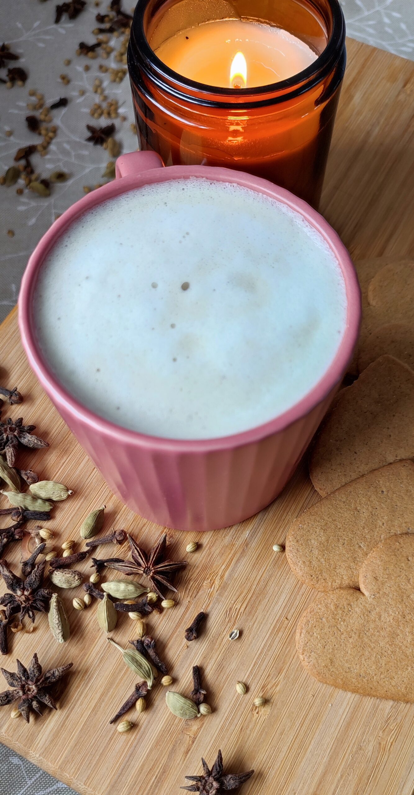 Masala Chai Latte in deep pink cup surounded with few hearts cookies on the right nd blend of whole spices on left and candle in a glass jar at the back