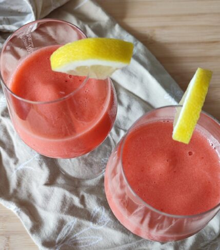 Pink Sgroppino in wine glasses garnished with quartered lemon wedges on the side on patterned taupe coloured fabric on a working surface