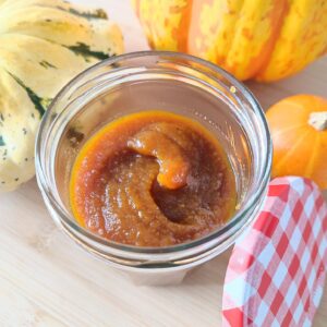 Pumpkin Spice Syrup in a small confiture jar with its red and white checkered patterned lid resting on the side vertically and three decorative pumpkins in the background