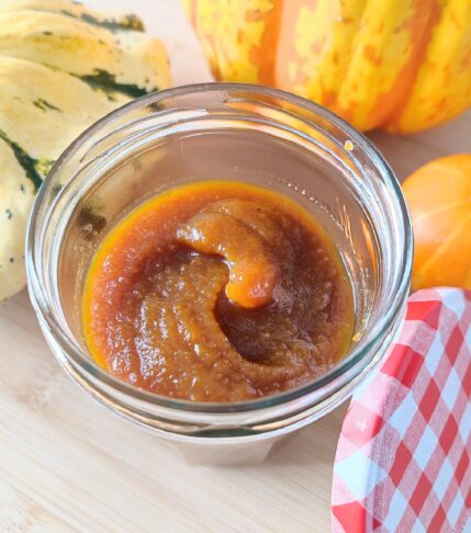 Pumpkin Spice Syrup in a small confiture jar with its red and white checkered patterned lid resting on the side vertically and three decorative pumpkins in the background