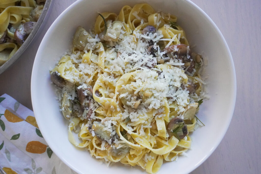 One Pan Lemon Butter Tagliatelle with Mushrooms & Artichokes in a large white bowl on a tea towel with lemon prints and the pan filled with the remaining pasta on the side