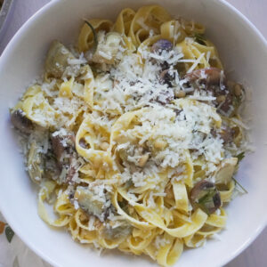 One Pan Lemon Butter Tagliatelle with Mushrooms & Artichokes in a large white bowl on a tea towel with lemon prints and the pan filled with the remaining pasta on the side