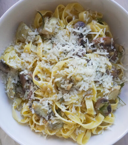 One Pan Lemon Butter Tagliatelle with Mushrooms & Artichokes in a large white bowl on a tea towel with lemon prints and the pan filled with the remaining pasta on the side