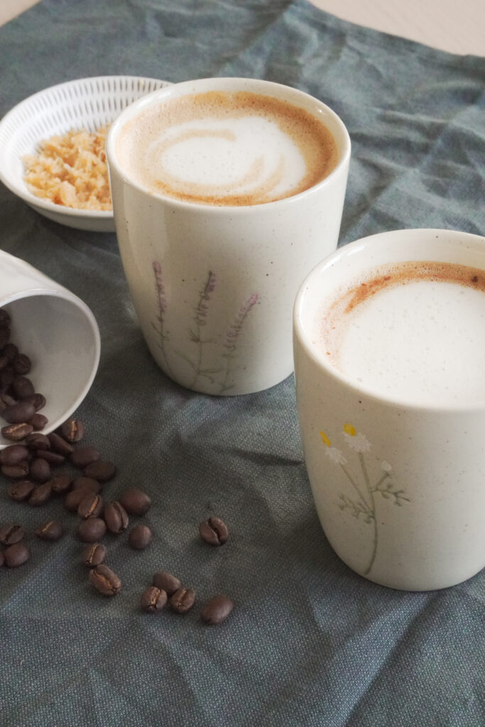 Dubai Chocolate Inspired Mocha in two cups with some coffee beans on the side and crunchy knafeh topping on the side on a dark green fabric from a side shot