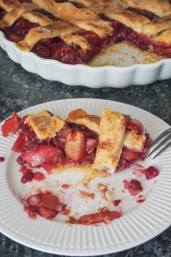 B roll 2: A slice of Apple Cherry Almond Pie half eaten on a white ribbed plate while the leftover pie is seen in background