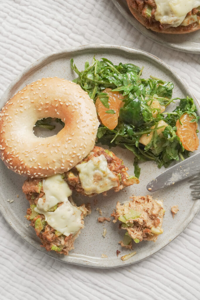 B roll 1: Top view of half eaten Airfryer Tuna Bagel Melt with a side of wild peach and mandarin arugula salad on modern ceramic plate and another airfryer tuna bagel melt in the background on textured beige table cloth