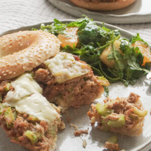 Half eaten Airfryer Tuna Bagel Melt with a side of wild peach and mandarin arugula salad on modern ceramic plate and another airfryer tuna bagel melt in the background on textured beige table cloth