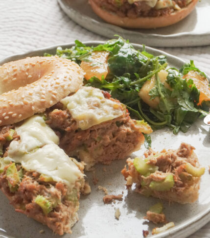 Half eaten Airfryer Tuna Bagel Melt with a side of wild peach and mandarin arugula salad on modern ceramic plate and another airfryer tuna bagel melt in the background on textured beige table cloth