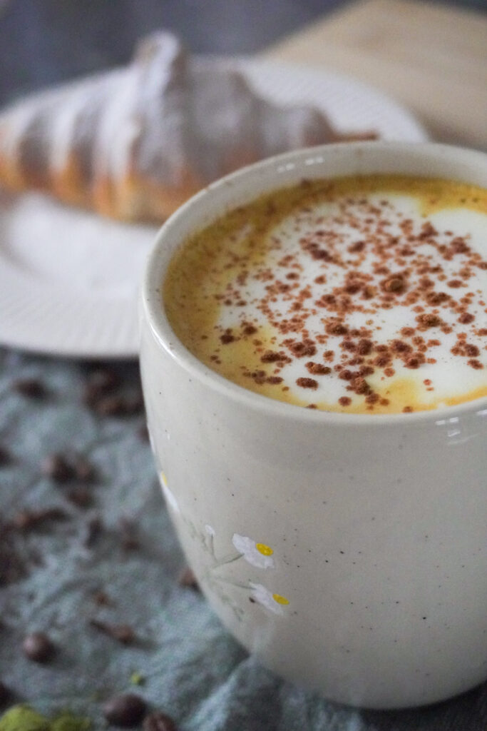 Spiced Pear & Turmeric Maple Latte with Soy Milk in a beige mug with a croissant with powdered sugar dusted on it on a white plate in the background and both are on a green cloth with spices, coffee beans, matcha powder and tea leaves scattered
