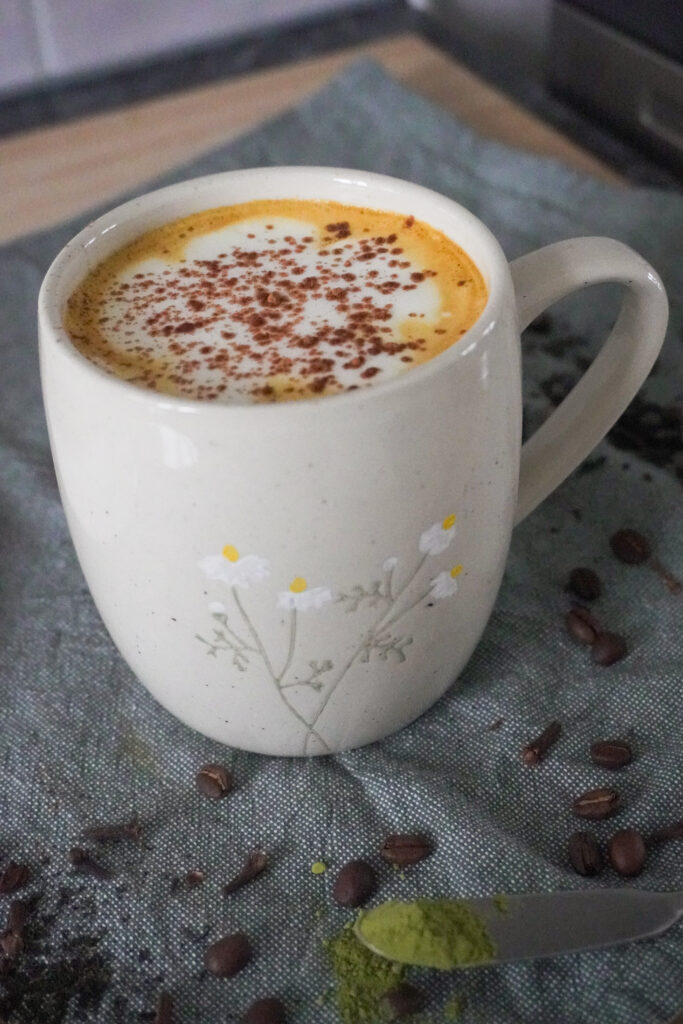 Spiced Pear & Turmeric Maple Latte with Soy Milk in a beige mug on green cloth with spices, coffee beans, matcha powder and tea leaves scattered